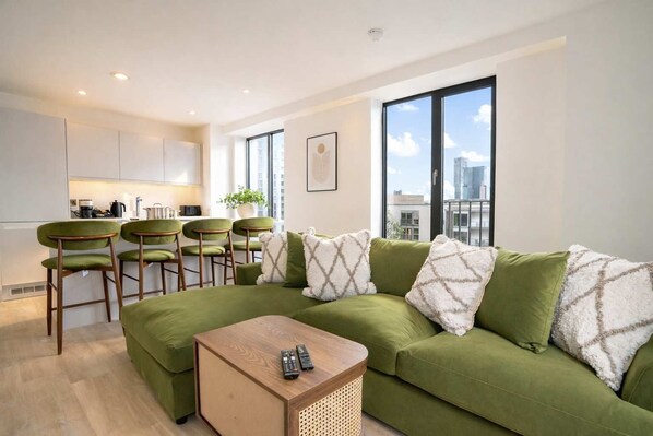 Un salon moderne avec un canapé vert, des fauteuils assortis, une table basse en bois et une vue sur la ville grâce à de grandes fenêtres.
