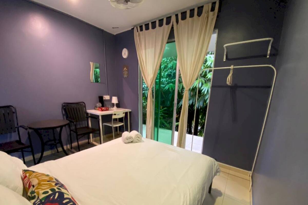 Un dormitorio con cama, escritorio y silla. Hay una ventana con cortinas y vistas al exterior con vegetación.