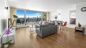 Een moderne woonkamer met een grijze bank, een eettafel met stoelen, een groot raam met uitzicht op de stad, en een bijzettafeltje met een plant.