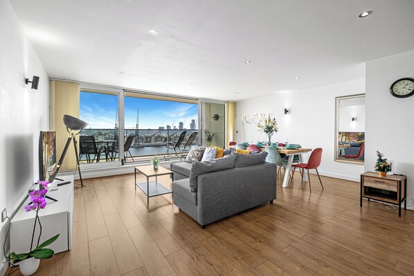 A modern living room with a grey sofa, a dining table with chairs, a large window with a city view, and a small side table with a plant.