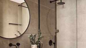 Ein modernes Badezimmer mit einem gebogenen Waschtisch, einem runden Spiegel, einer Dusche mit Handbrause und einer kleinen Pflanze auf dem Waschtisch.