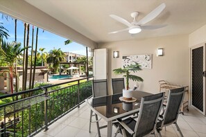 A balcony with a glass table, chairs, a ceiling fan, a potted plant, and a view of a swimming pool and buildings. - Tropical Garden Retreat with 9 Pools (Westcourt)
