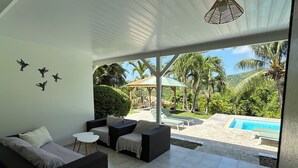 A modern room with a sofa, a small table, and a view of a pool and garden. - LA VILLA LES 4 COLIBRIS, 3 ch, piscine avec une vue imprenable sur la montagne  (LE VAUCLIN)