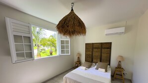 A bedroom with a bed, two bedside tables, a hanging light fixture, a window with shutters, and an air conditioning unit. - LA VILLA LES 4 COLIBRIS, 3 ch, piscine avec une vue imprenable sur la montagne  (LE VAUCLIN)