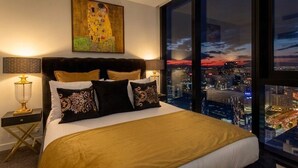 Une chambre d’hôtel avec un grand lit, une vue sur la ville, un tableau accroché au mur et un ciel étoilé visible par la fenêtre.