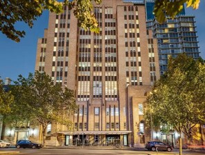 Un edificio de varios pisos con una entrada central, ventanas amplias y un estacionamiento al frente.