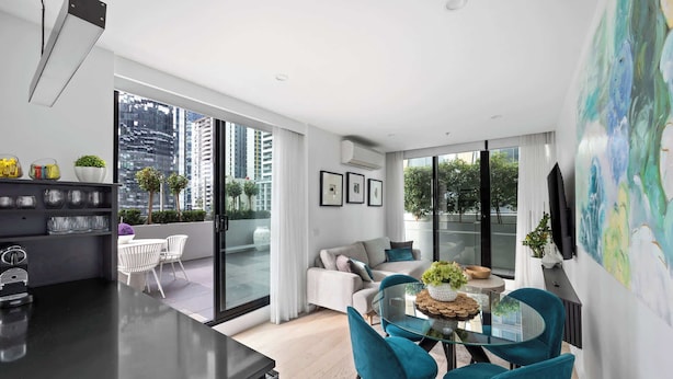 A modern living area with a glass dining table, teal chairs, a sofa, and a large window offering a city view. - Huge private wrap-around terrace near Crown Casino (Southbank)