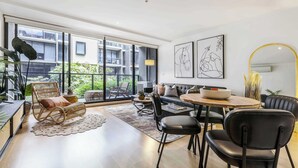 A modern living room with a sofa, chairs, a dining table, and a large window with a view of a building and greenery. - Tropical courtyard sanctuary near Glenferrie Rd (Hawthorn)
