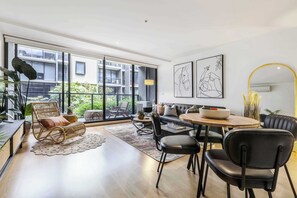 Una sala de estar moderna con un sofá, sillas, una mesa de comedor y un ventanal con vistas a un edificio y follaje.