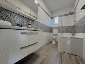 Ein modernes Badezimmer mit Toilette, Waschbecken und Dusche. Es verfügt über eine geflieste Spritzschutzwand, Holzboden und weiße Schränke.