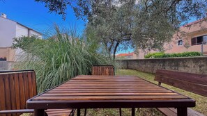 Eine Terrassengruppe im Freien mit Holztisch und Bänken, umgeben von hohem Gras und Bäumen.