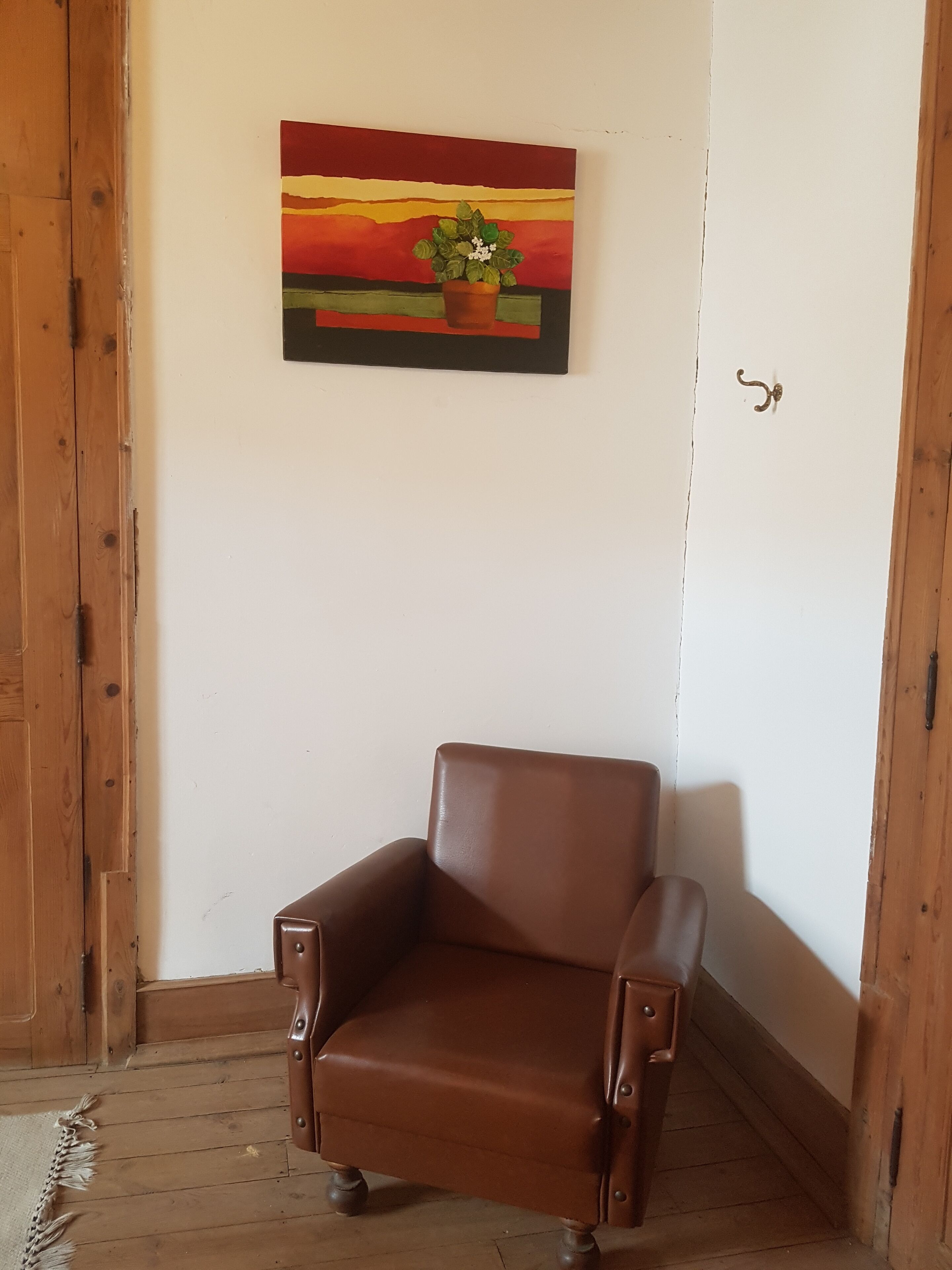Une pièce d’angle avec un fauteuil en cuir marron, un plancher en bois et un tableau représentant un coucher de soleil, ainsi qu’une plante accrochée au mur.