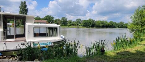 A houseboat docked by a calm lake with a grassy shore and trees in the background.