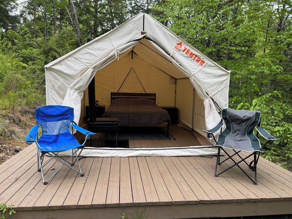 Una carpa con una cama, dos sillas y una pequeña mesa sobre una terraza de madera rodeada de árboles.