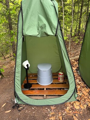 Una carpa verde con un inodoro y un bote de basura en su interior, rodeada de un bosque.