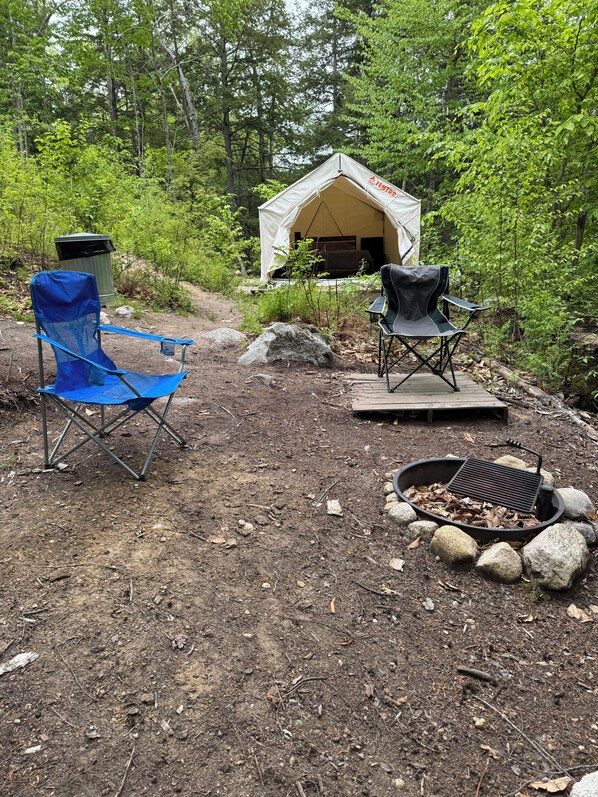 A tent with a sleeping area, two camping chairs, a fire pit, and a trash can in a forest setting.