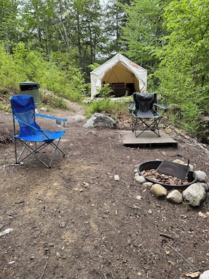 Una carpa con zona de dormir, dos sillas de camping, un brasero y un bote de basura en un entorno boscoso.