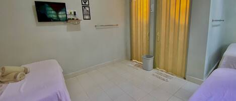 A hotel room with two beds, a TV, a small shelf with toiletries, a trash bin, and a window with curtains.