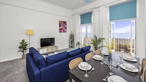 A modern living room with a blue sofa, dining table set for four, and a TV on a wooden cabinet. - The Cheeky Chappie By My Getaways (Brighton)