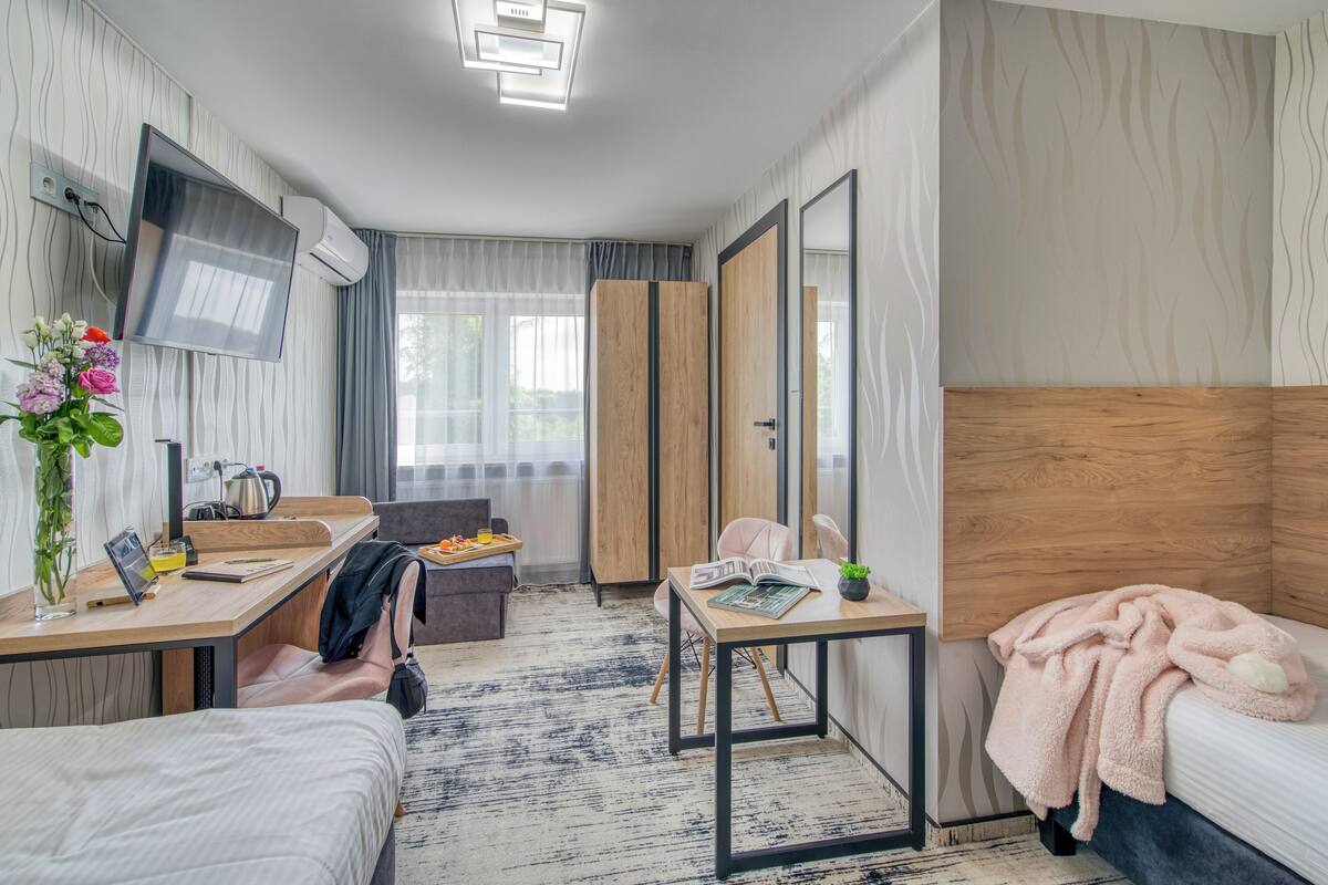 A modern hotel room with a bed, a desk with a chair, a TV, and a large mirror.