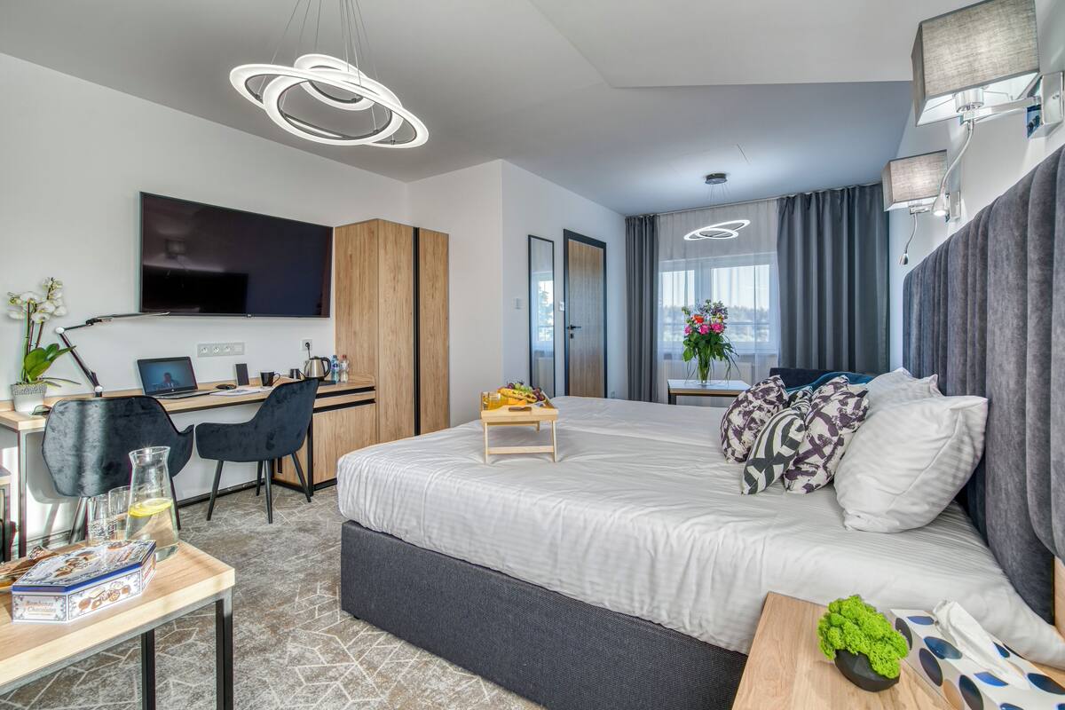 A modern hotel room with a large bed, a desk with a laptop, a TV, and a view of the city.