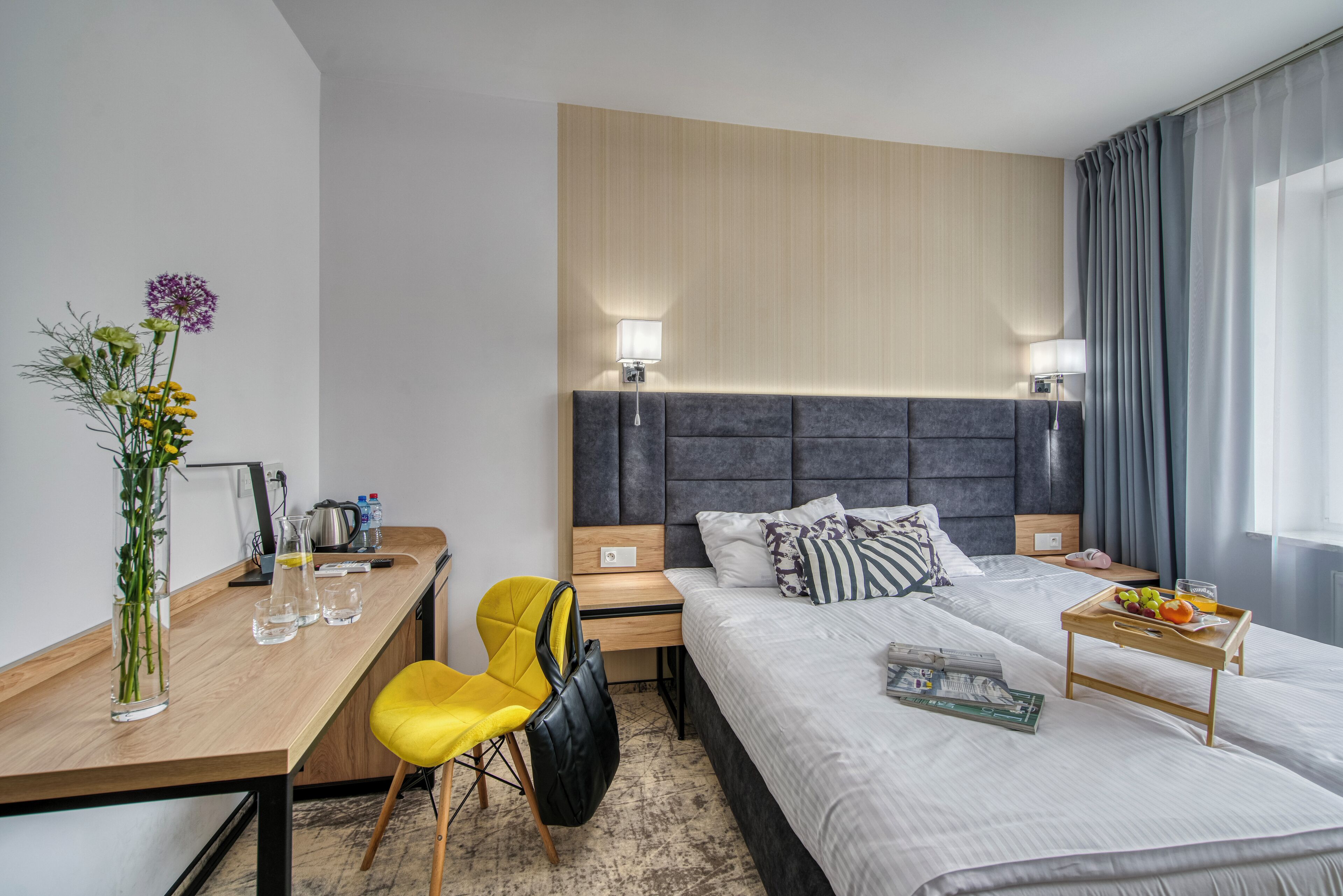 A modern hotel room with a bed, bedside tables, a desk, a chair, and a vase with flowers.