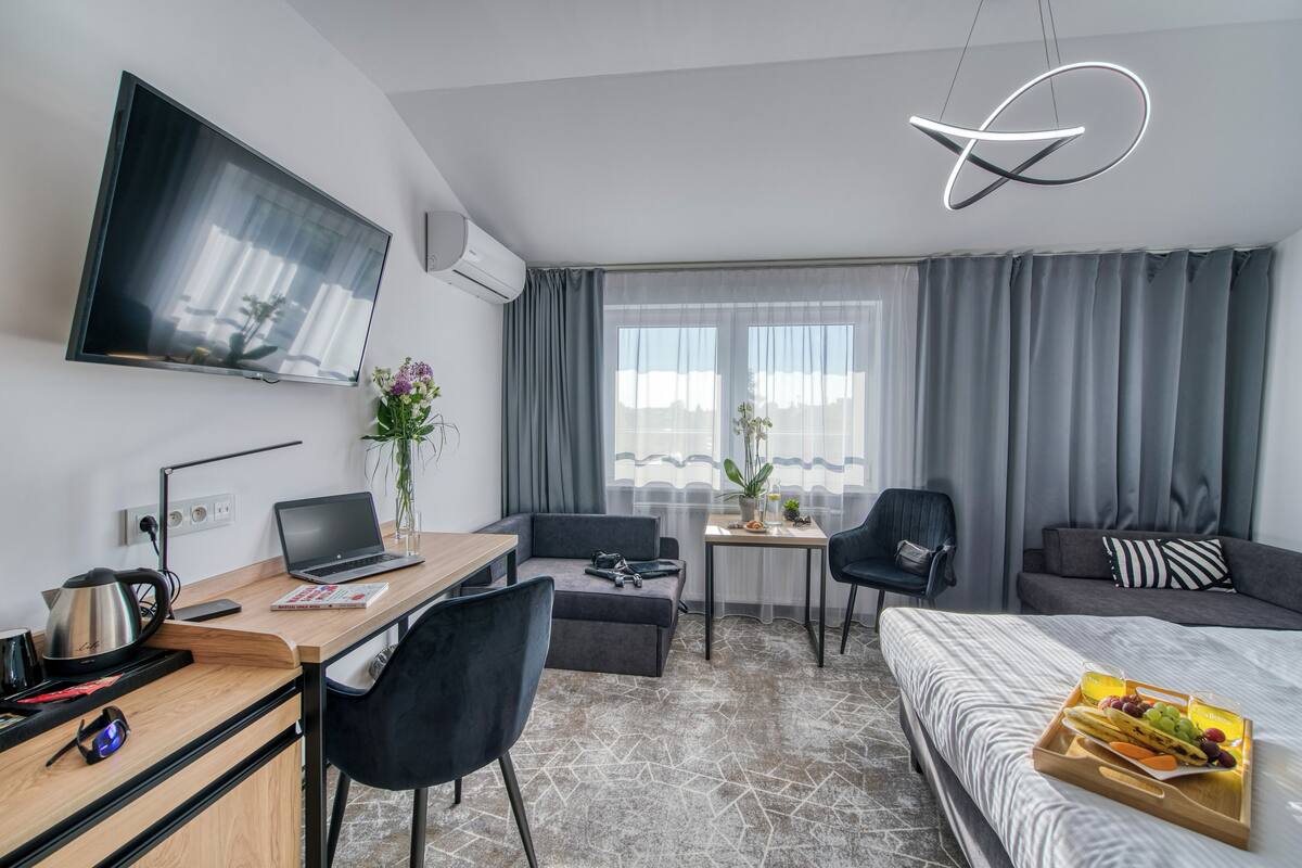 A modern hotel room with a flat-screen TV, a desk with a laptop, a sofa, a bed, and a dining table with a tray of fruit.