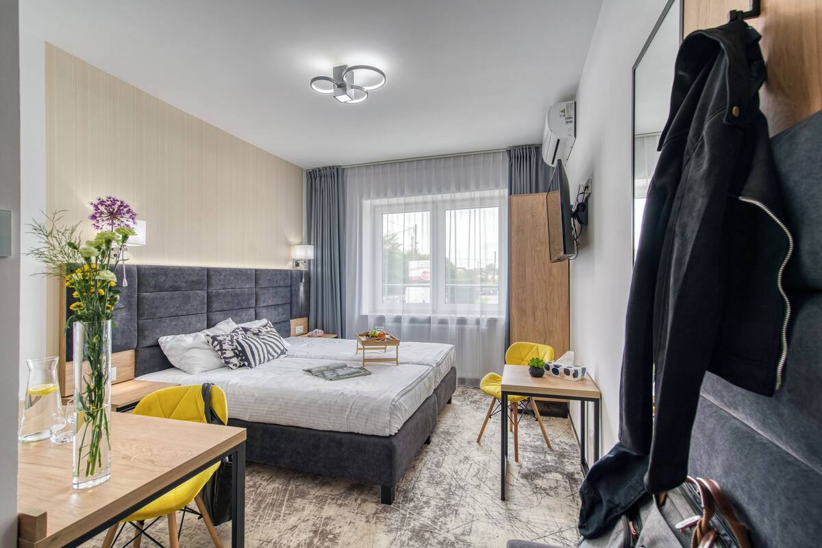A modern hotel room with a large bed, a desk, a chair, a small table, a vase with flowers, and a coat rack.