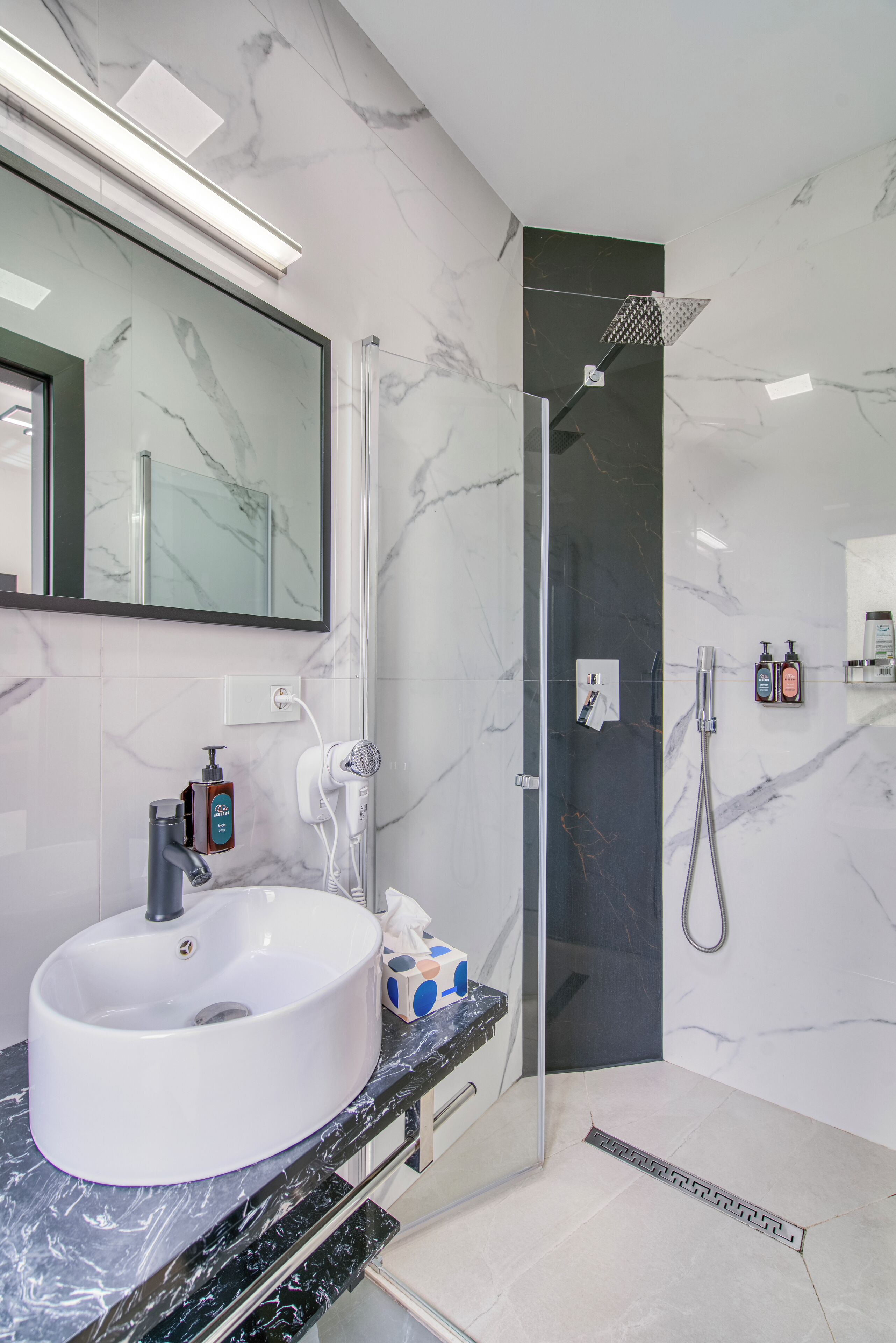 Een moderne badkamer met een witte wastafel, een grote spiegel, een douche met glazen wanden en marmeren wanden.