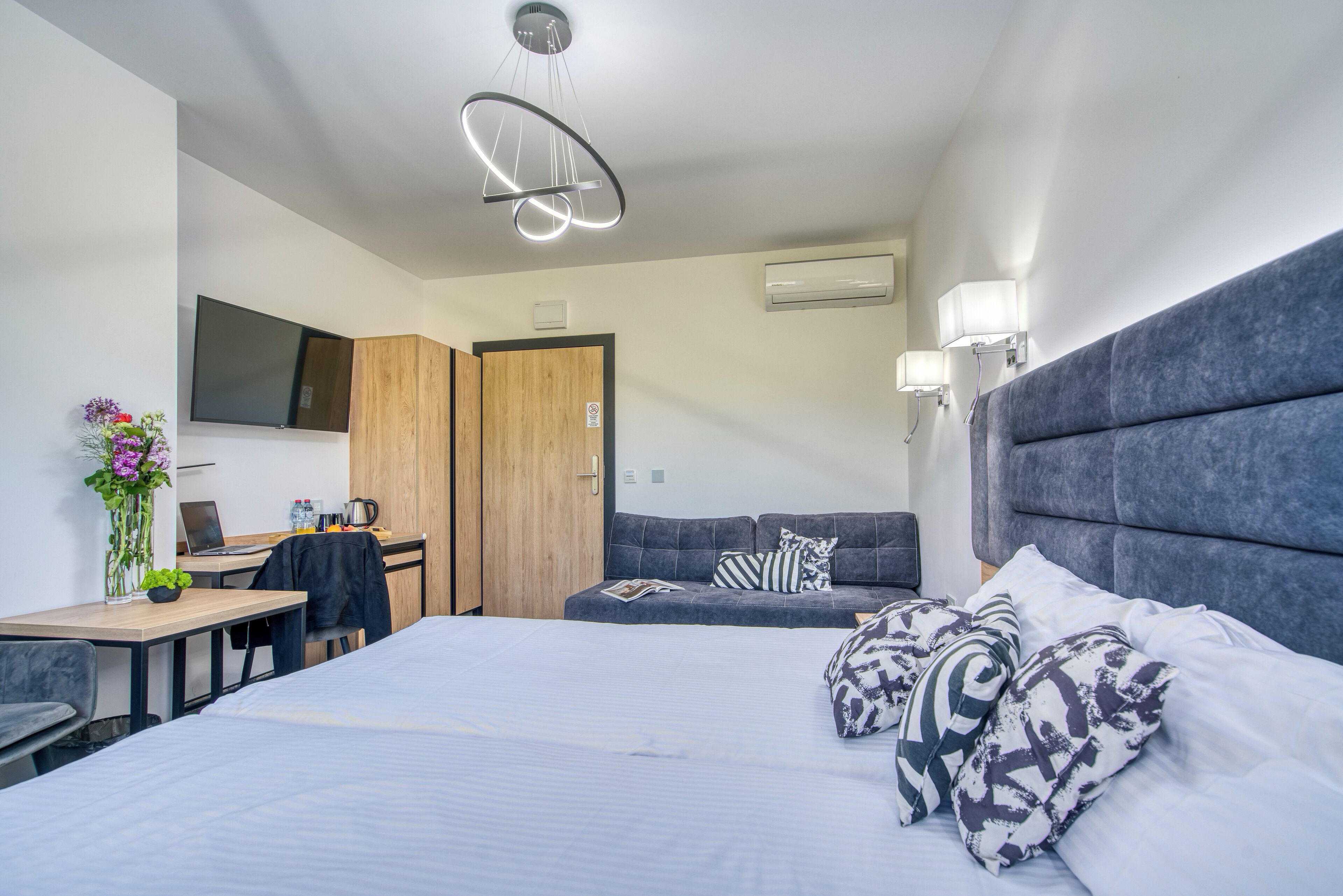 A modern hotel room with a large bed, a desk with a laptop, a sofa, and a TV mounted on the wall.