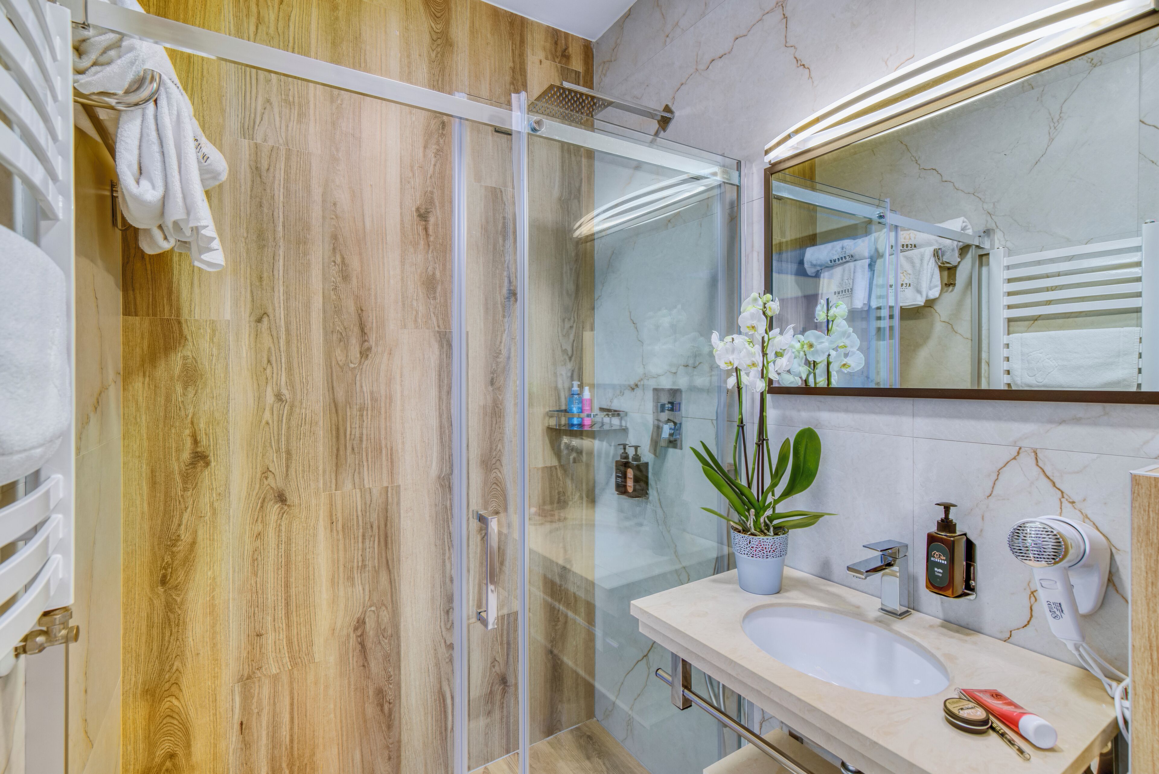 Bany modern amb paret de fusta, pantalla de dutxa de vidre, un lavabo amb mirall i una orquídia en test.