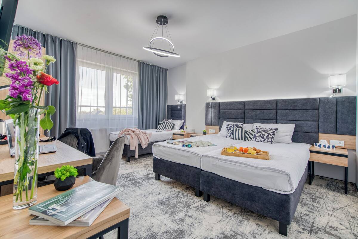 A modern hotel room with a large bed, a desk with a laptop, a vase with flowers, and a view of the outdoors.
