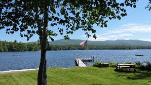 A lakeside view with a dock, grassy area, picnic table, and an American flag. - Quintessential Winnipesaukee Lake Cottage Sleeps 8 with Views and Dock (Moultonborough)