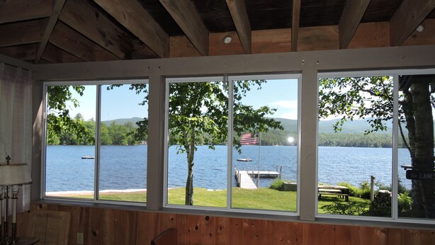 A cabin with large windows overlooking a lake, wooden interior, and a dock visible through the windows. - Quintessential Winnipesaukee Lake Cottage Sleeps 8 with Views and Dock (Moultonborough)