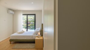 A bedroom with a bed, a nightstand, a lamp, a window with a view of greenery, and an air conditioning unit. - Lajinha do Ave (Braga)