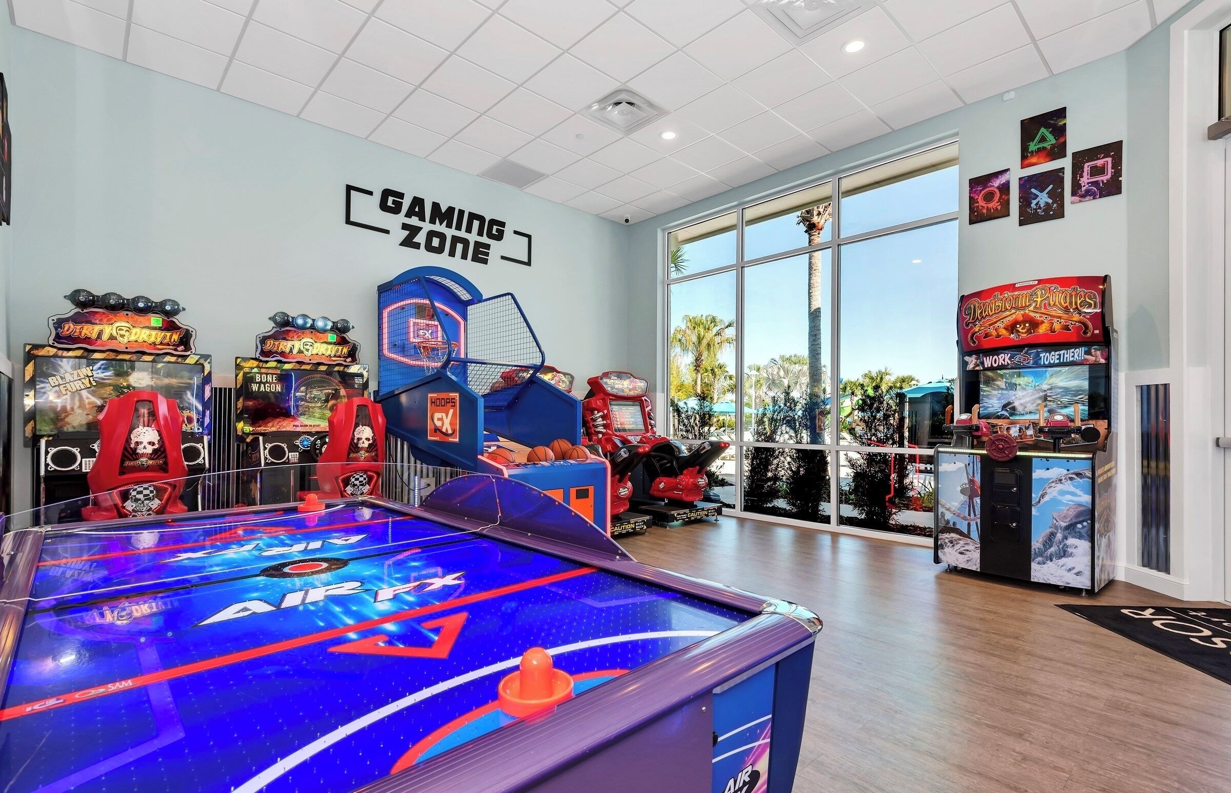 A gaming zone with various arcade machines, a pool table, and large windows offering a view of palm trees.