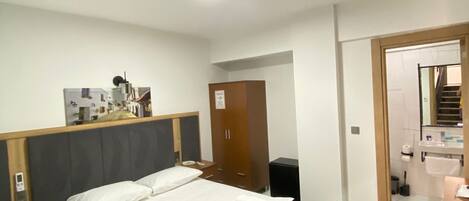 A hotel room with a large bed, a wooden headboard, a bedside table, a wardrobe, and a bathroom visible through an open door.