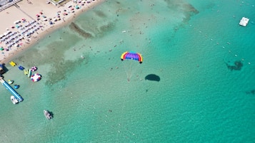 An aerial view of a beach with clear turquoise water, a parasailing activity, and beach umbrellas.