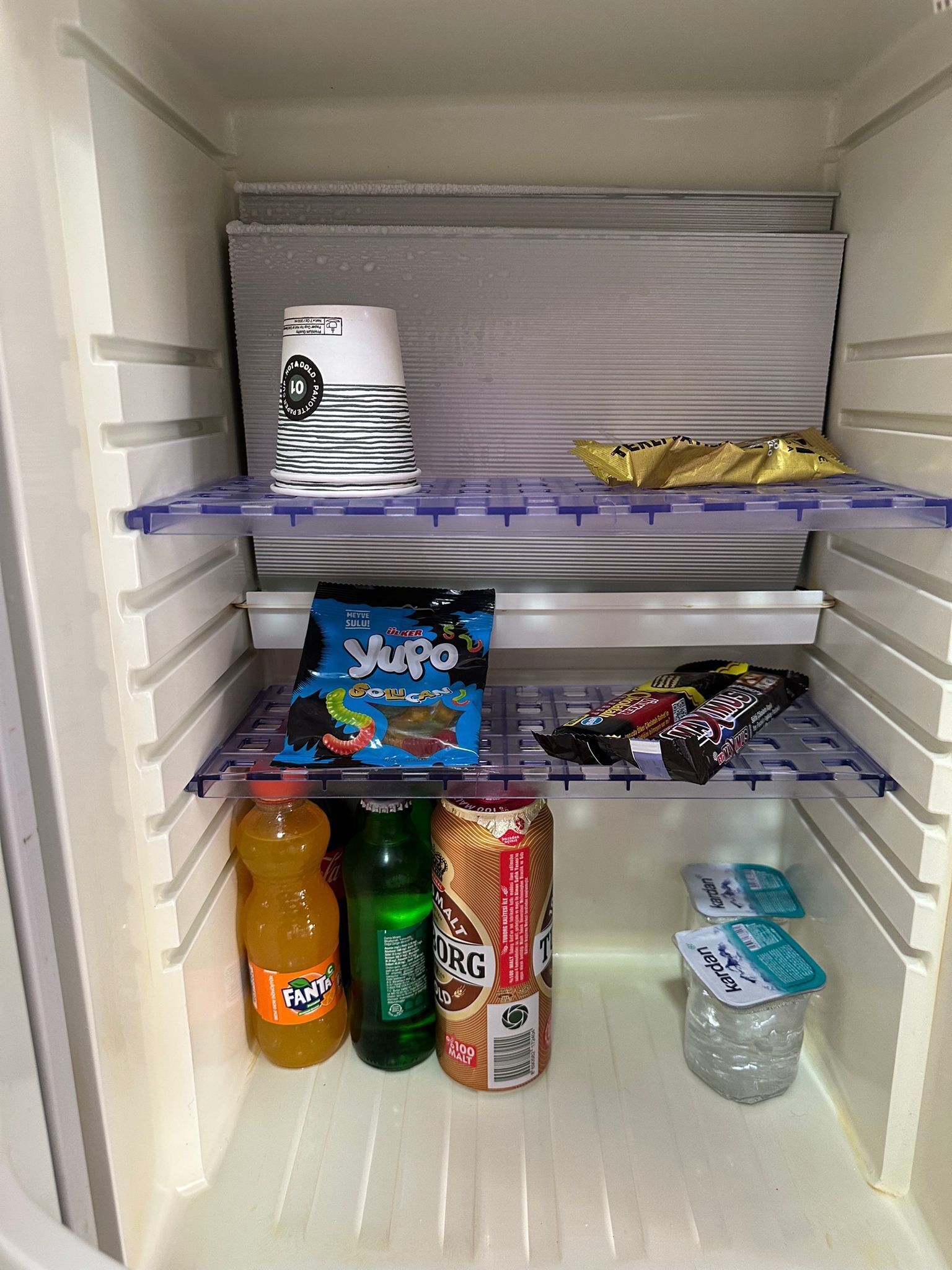 Un refrigerador lleno de diversos alimentos, incluyendo una botella de Fanta, un frasco verde, una lata de sal, una caja de Yupo y una barra de chocolate.