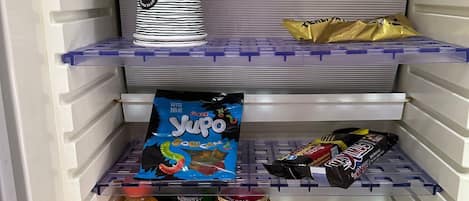 A refrigerator stocked with various food items including a bottle of Fanta, a green bottle, a can of salt, a box of Yupo, and a pack of chocolate bars.