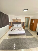 Habitación de hotel con una cama grande, un armario de madera, un baño visible a través de una puerta abierta y una mesita con una lámpara.