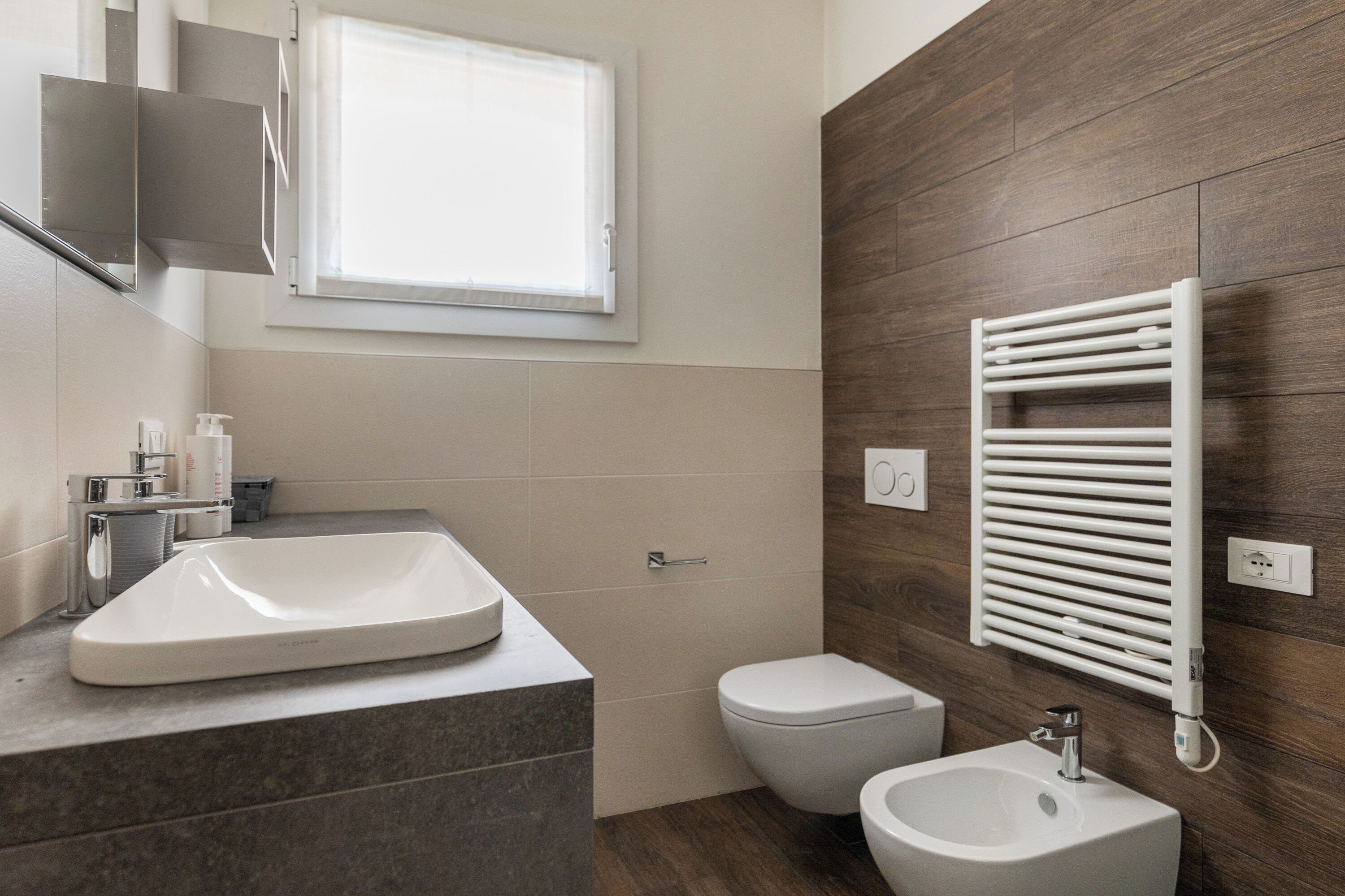 Une salle de bain moderne dotée d’un grand lavabo, de toilettes et d’un bidet, avec un mur en bois et une fenêtre.