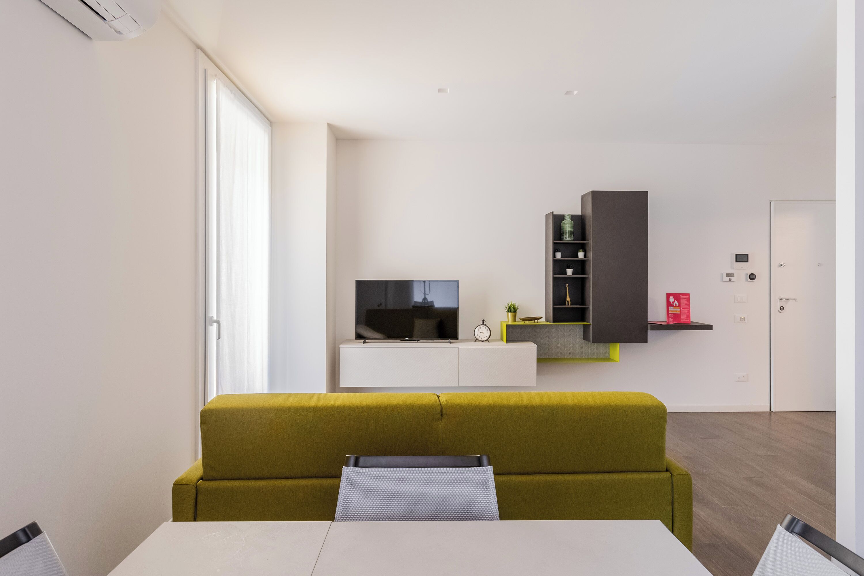 A modern living room with a sofa, a dining table, and a kitchen area with a TV, cabinets, and a door.