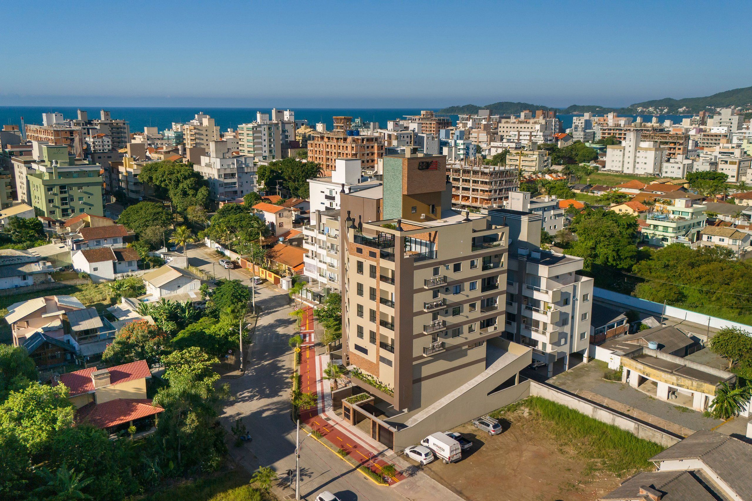 A high-rise building with multiple floors, surrounded by other residential buildings, a road with vehicles, and a view of the sea in the background.