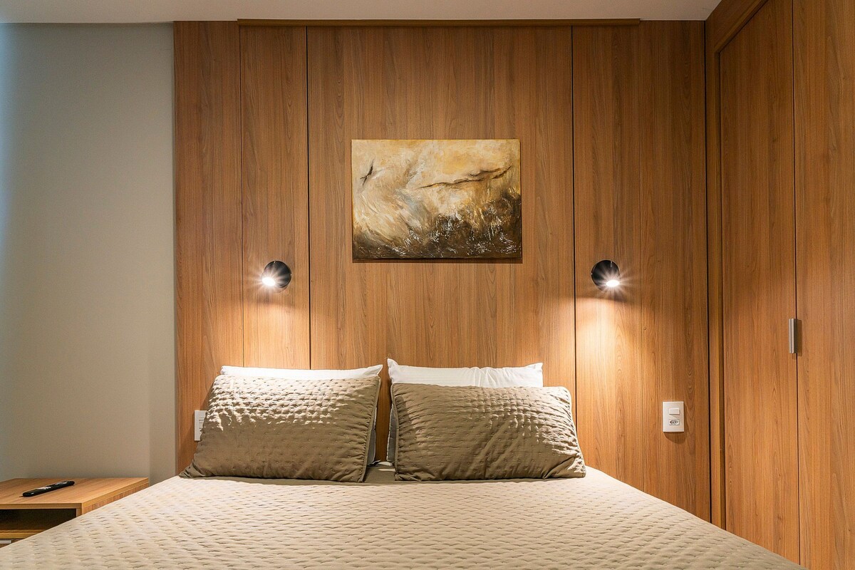 Un dormitorio con cabecera de madera, cama con ropa de cama color crema, dos lámparas de lectura fijadas a la pared y un cuadro sobre la cama.
