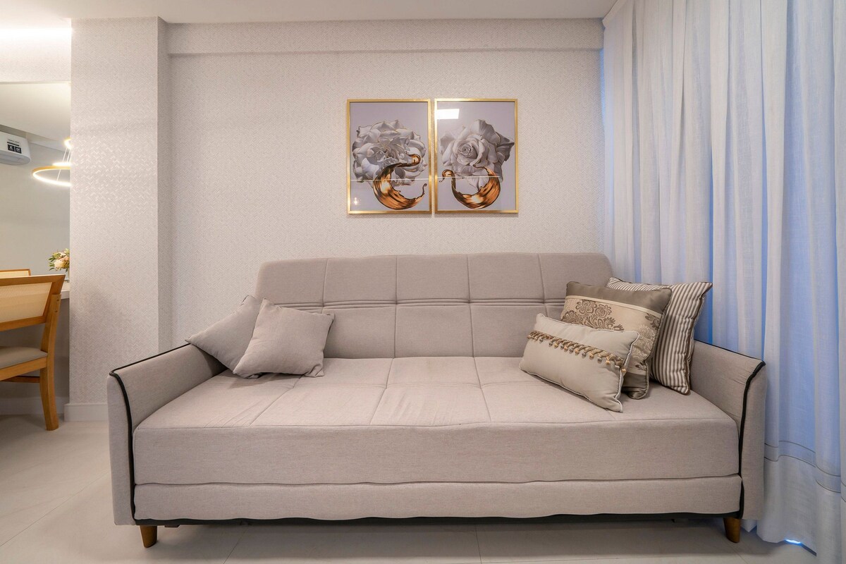 Una sala de estar con un sofá color beige, dos cuadros enmarcados con motivos florales y un comedor con una mesa y sillas de madera.