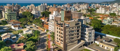 Un edificio de gran altura con varios pisos, rodeado de otros edificios residenciales, una calle con vehículos y una vista al mar en el fondo.