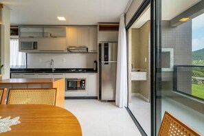 A modern kitchen with built-in appliances, a dining area with a wooden table and chairs, and a balcony with a view of greenery. - Las Vegas  (Bombinhas)