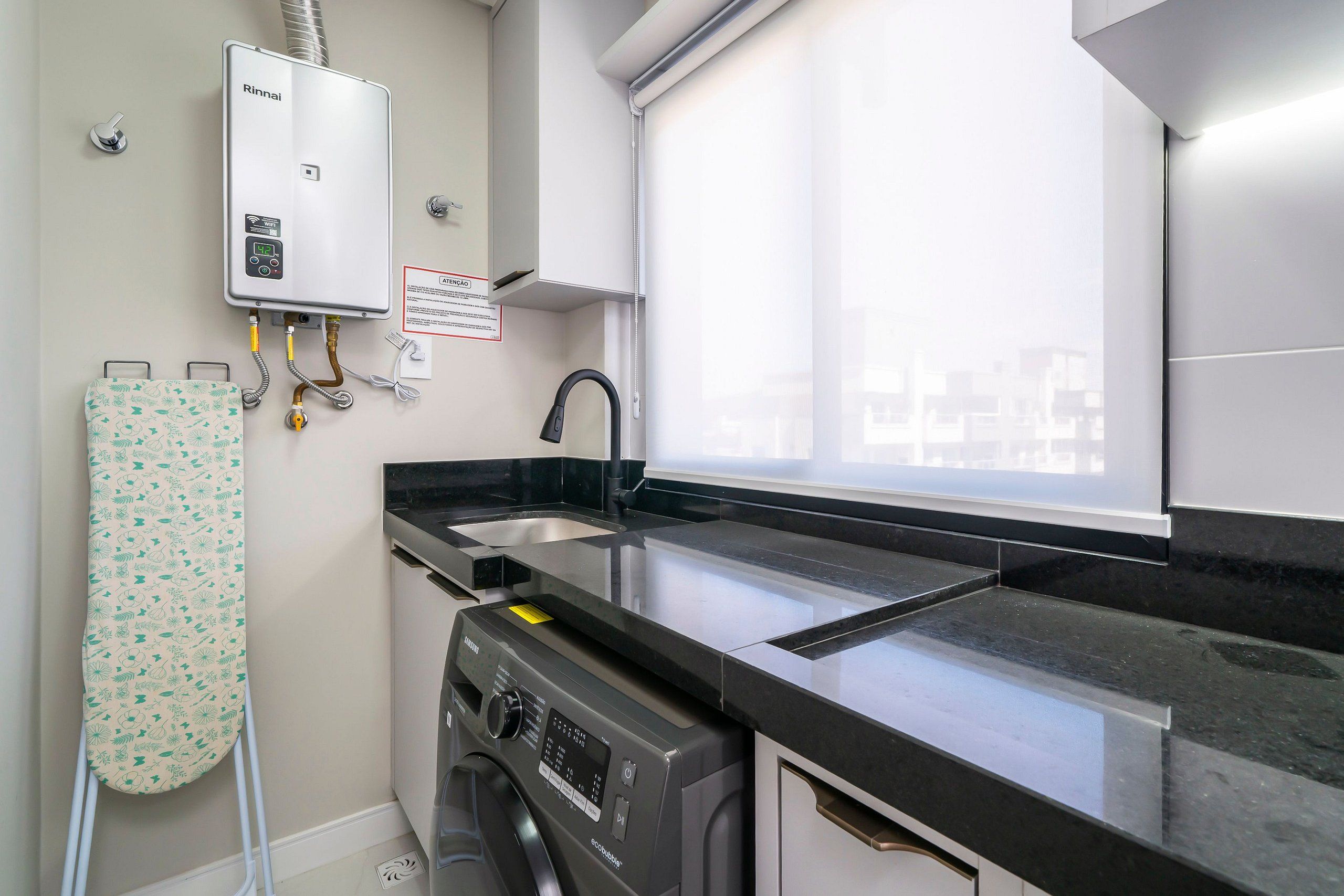 A compact kitchen with a Rinnai water heater, a washing machine, and a window with a city view.