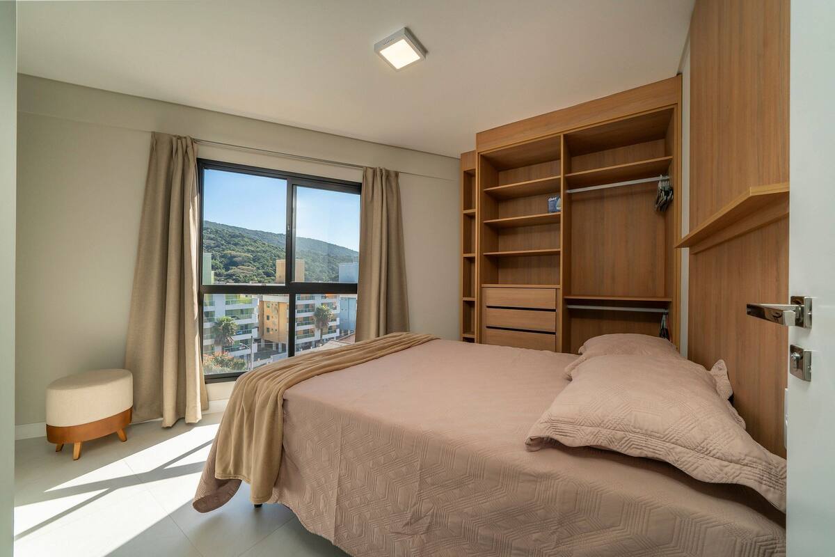 Une chambre avec un grand lit, une fenêtre avec des rideaux, une armoire en bois et une vue sur un immeuble et des collines.